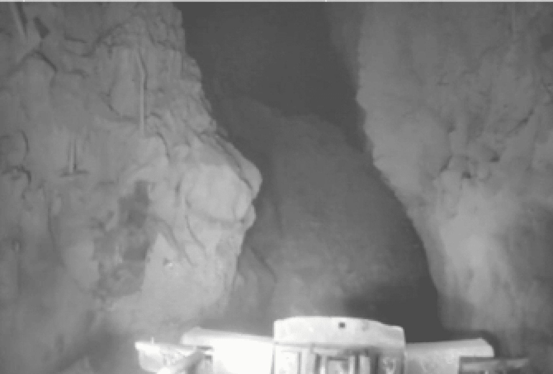 Grainy grayscale CCTV feed from an underground mine showing a bogger bucket with minimal visibility